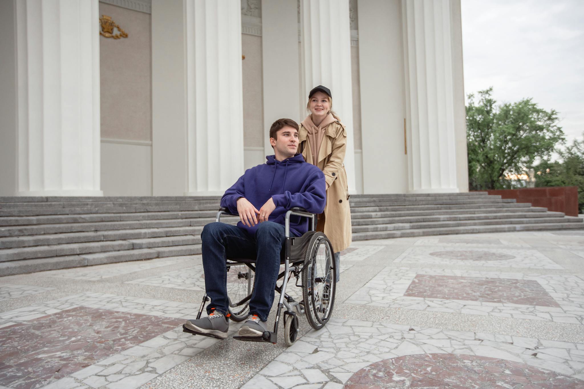 Woman assisting a man in a wheelchair outside a building with classical columns.